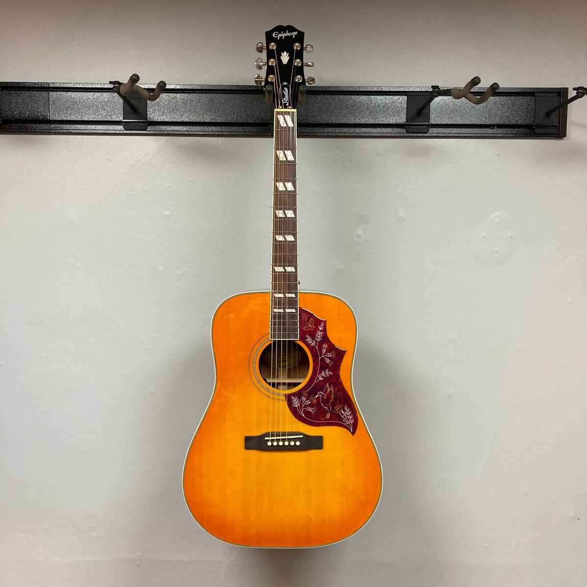 An Epiphone Hummingbird Studio Faded Cherry Sunburst acoustic-electric guitar with decorative pickguard hangs on a black wall rack against a white wall, featuring a built-in Fishman Sonitone pickup. Includes gigbag.
