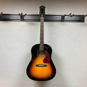 An Epiphone J-45 Studio Vintage Sunburst w/Gigbag six-string acoustic guitar hangs on a wall-mounted rack against a white wall.
