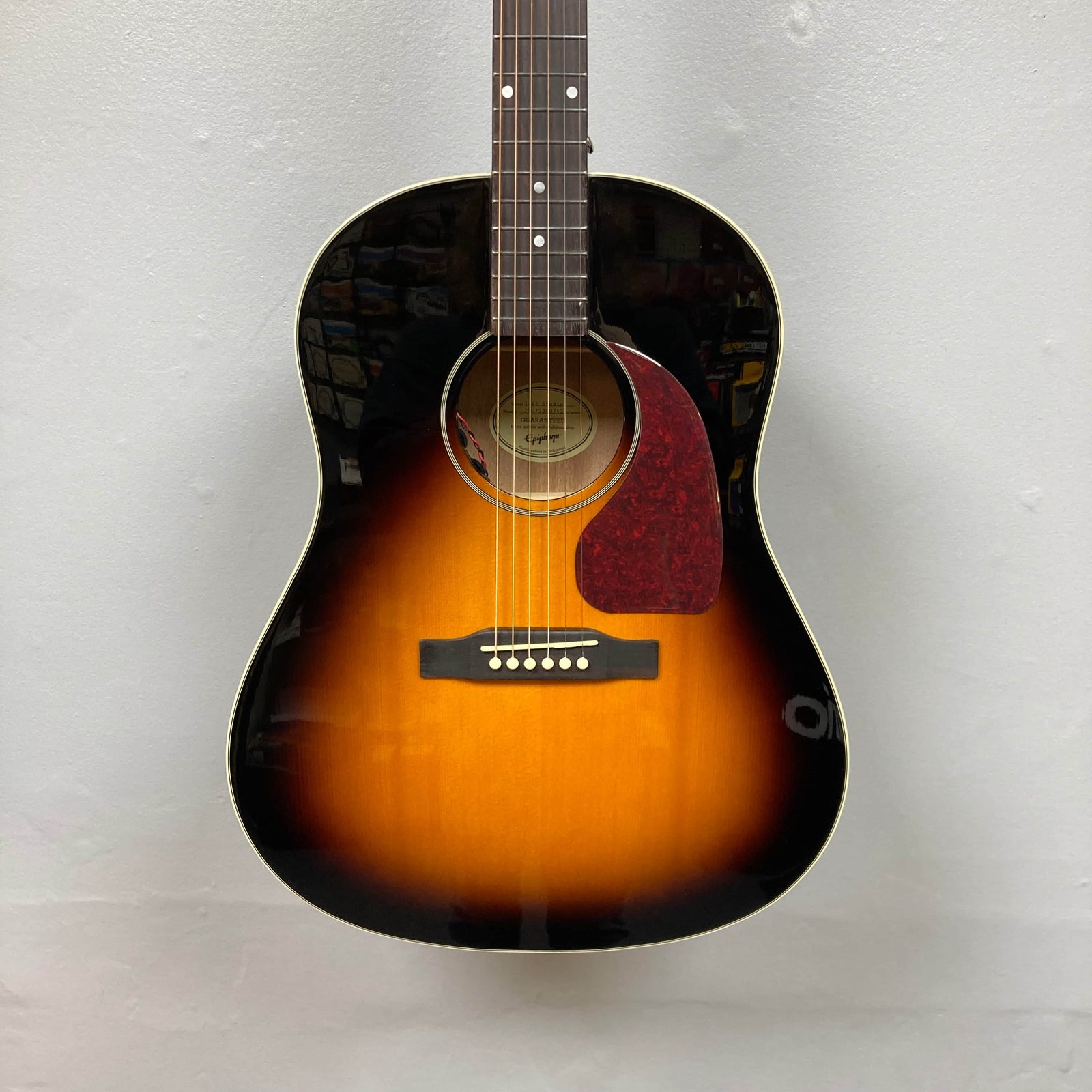 An Epiphone J-45 Studio Vintage Sunburst acoustic guitar with six strings, solid spruce top, and reddish-brown pickguard is shown against a plain white wall. Includes gigbag. Brand: Epiphone. An Epiphone J-45 Studio Vintage Sunburst acoustic guitar with six strings, solid spruce top, and reddish-brown pickguard is shown against a plain white wall. Includes gigbag. Brand: Epiphone.