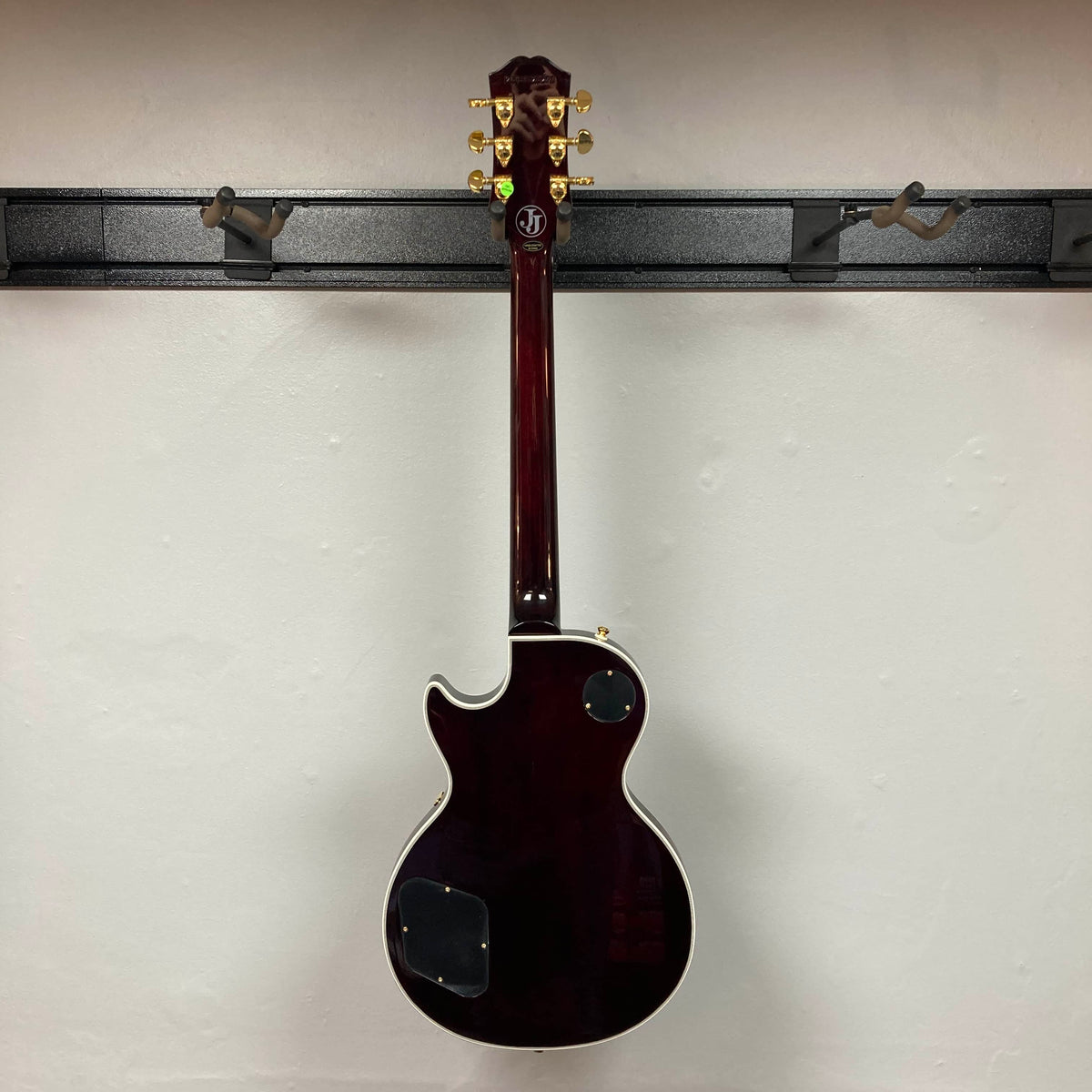 An Epiphone Jerry Cantrell Wino Les Paul Custom in Wine Red with gold tuning pegs and white trim, shown from the back, hangs on a wall-mounted rack. Case included.