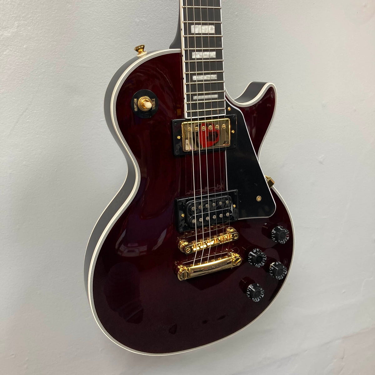 An Epiphone Jerry Cantrell Wino Les Paul Custom in Wine Red with gold hardware, black pickguard, and white trim hangs on a plain white wall.