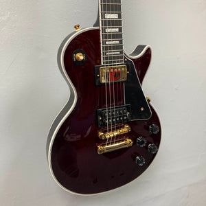 An Epiphone Jerry Cantrell Wino Les Paul Custom in Wine Red with gold hardware, black pickguard, and white trim hangs on a plain white wall.
