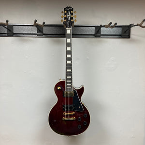 Epiphone Jerry Cantrell Wino Les Paul Custom Wine Red, 6-string electric guitar on wall hook, featuring gold hardware and custom design, includes hardshell case.