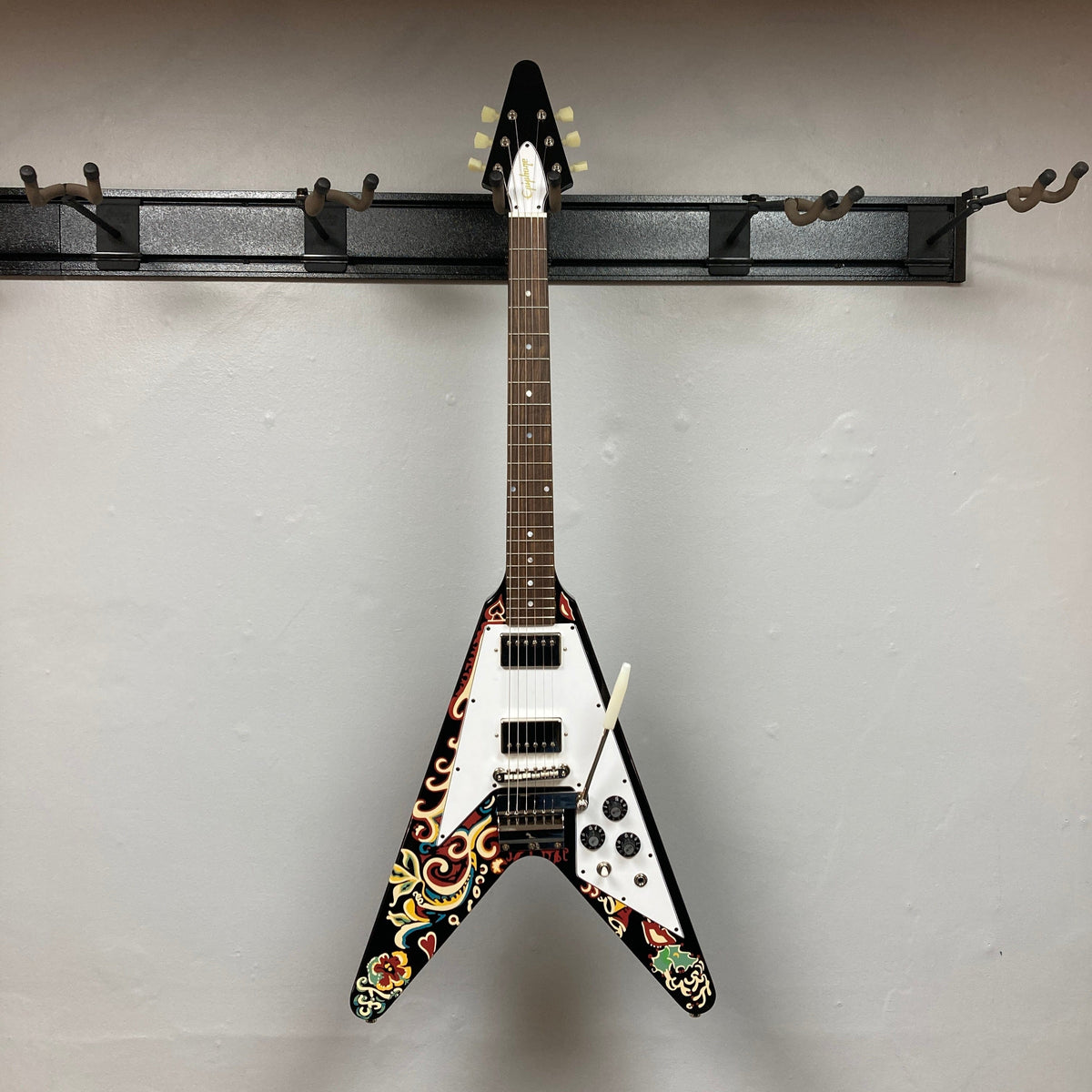 Epiphone Jimi Hendrix Love Drops Flying V guitar, on a wall hook, showcasing its iconic psychedelic design. Includes a hard case for protection.