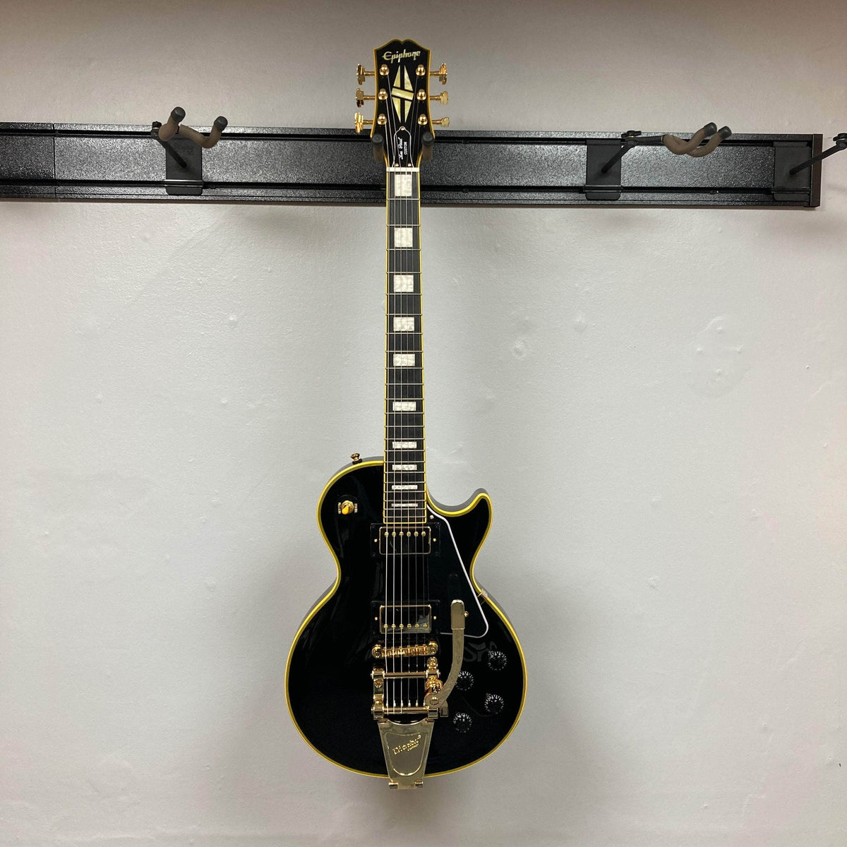 Epiphone Joe Bonamassa 59 Les Paul Custom Bigsby, featuring a glossy finish, block inlays, gold hardware, and a Bigsby tailpiece, mounted on a wall hanger.