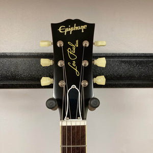 Close-up of an Epiphone Les Paul 1957 Goldtop Reissue Blem headstock, featuring six tuning pegs with white keys and visible logo, as it hangs on a wall-mounted holder. Model comes with case.