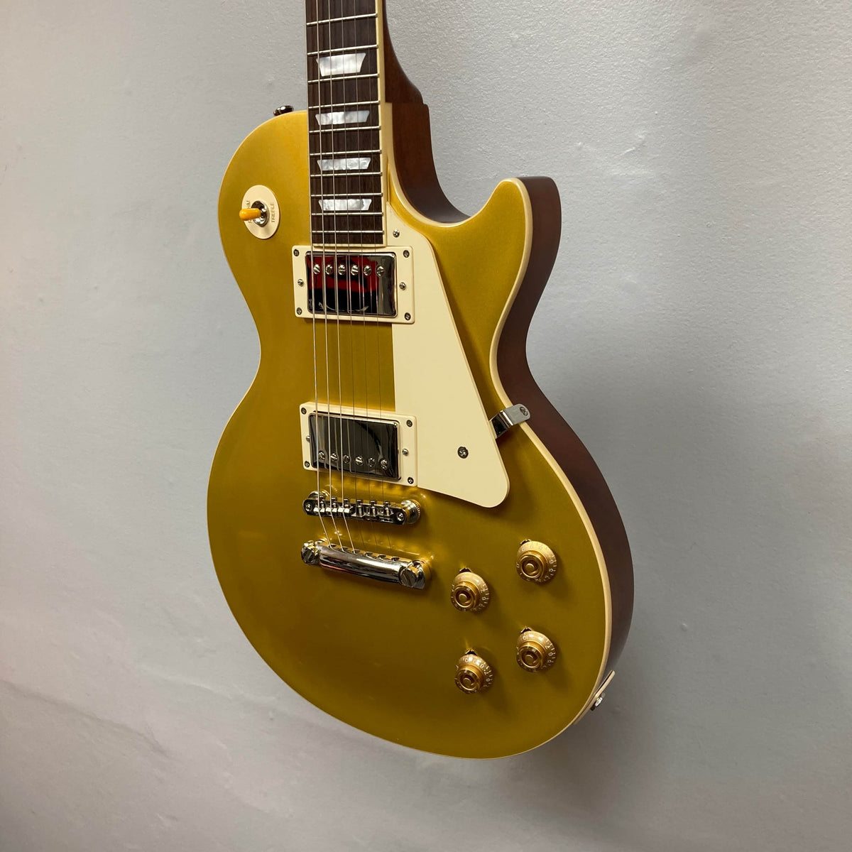 Epiphone Les Paul 1957 Les Paul Goldtop Reissue Blem w/Case—a gold electric guitar with white pickguard, four control knobs, and two humbucker pickups—hangs elegantly on a light grey wall.