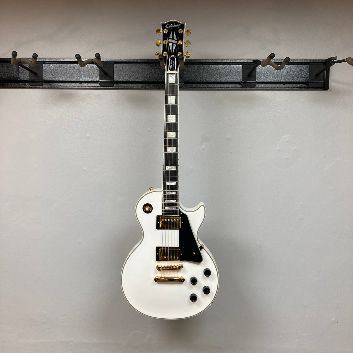 Epiphone Les Paul Custom Inspired by Gibson, Alpine White, displayed on a wall mount, featuring gold hardware, ebony fretboard, and mother-of-pearl inlays; includes hardshell case.