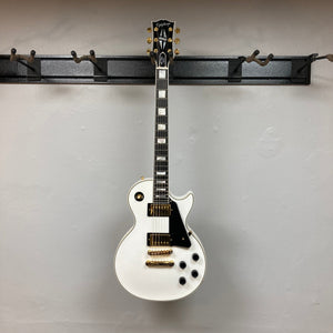 Epiphone Les Paul Custom Inspired by Gibson, Alpine White, displayed on a wall mount, featuring gold hardware, ebony fretboard, and mother-of-pearl inlays; includes hardshell case.