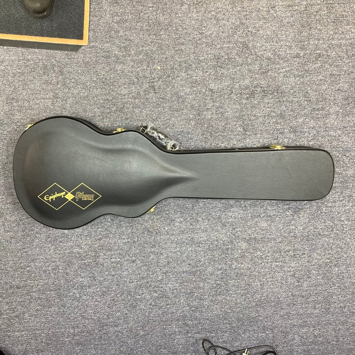 Epiphone Les Paul Custom guitar in black hardshell case on carpet, showcasing the Inspired by Gibson Custom logo.