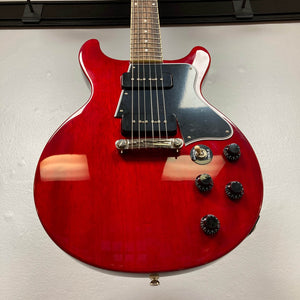 The Epiphone Les Paul Special Double Cut Cherry Red w/Gigbag electric guitar features two black pickups, four control knobs, and a three-way switch, inspired by Gibson, and is shown hanging on a white wall.