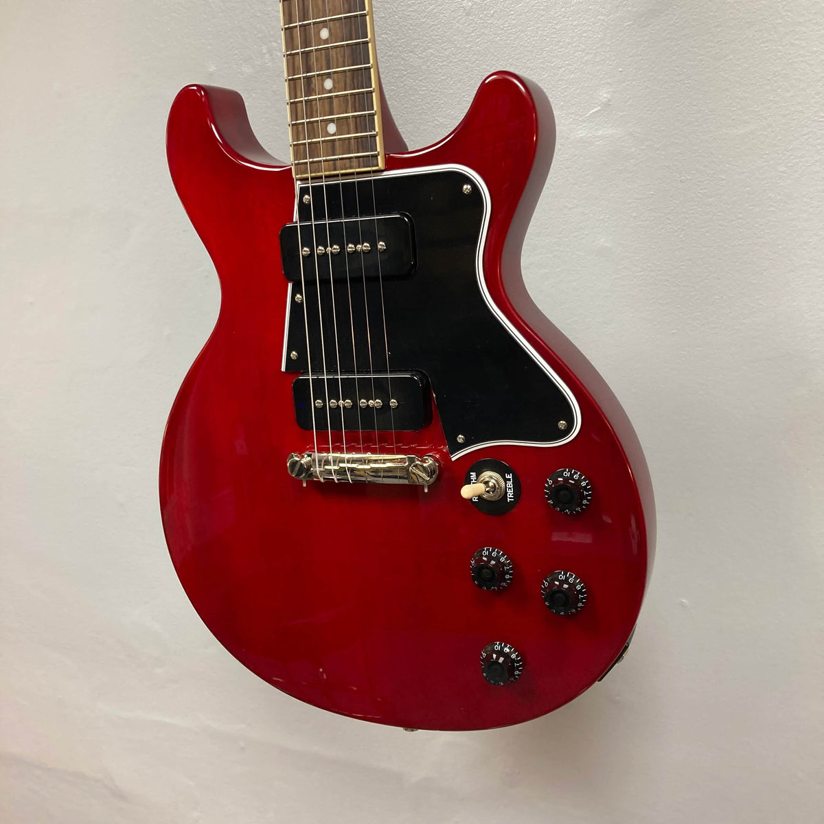 An Epiphone Les Paul Special Double Cut Cherry Red electric guitar with black pickguard, two pickups, four control knobs, and rosewood fretboard hangs on a white wall. Includes gigbag. Inspired by classic Gibson design.