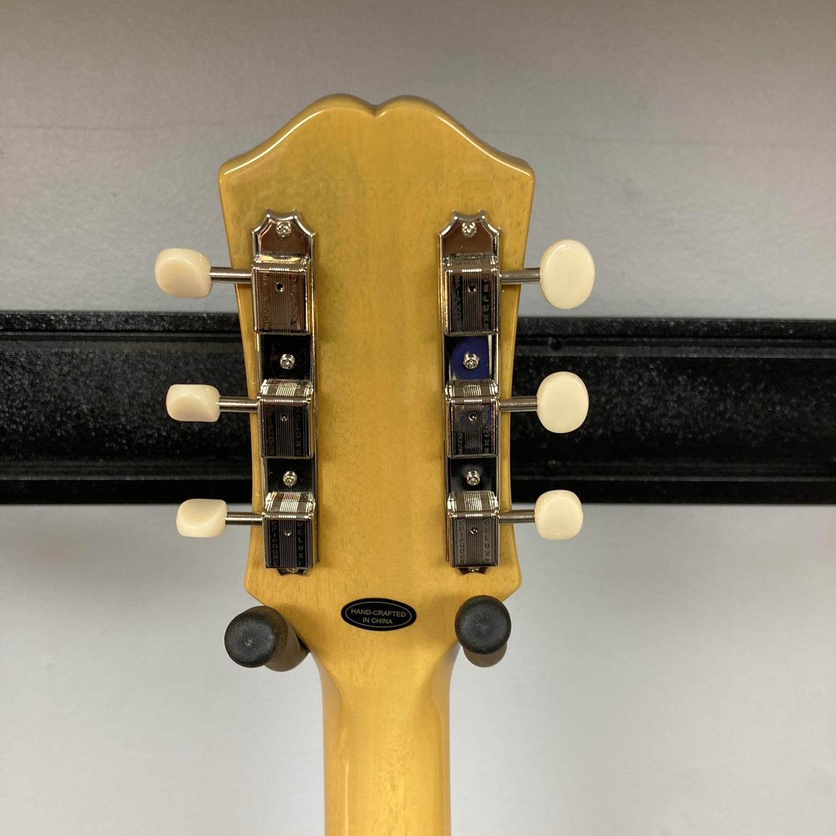 The back of an Epiphone Les Paul Special Double Cut TV Yellow w/Gigbag headstock with six tuning pegs is shown on a wall hanger against a plain background.