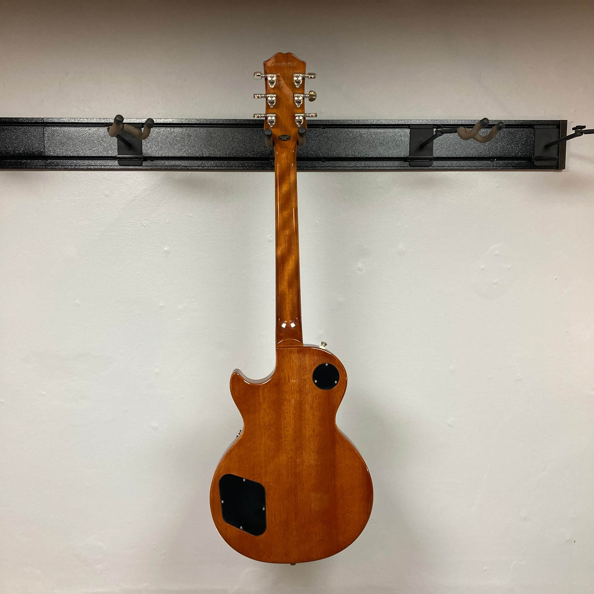 An Epiphone Les Paul Standard 60s Ebony w/Gigbag hangs with its back forward on a black metal rack, showing two control cavity covers.