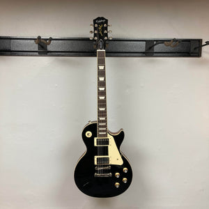 An Epiphone Les Paul Standard 60s Ebony w/Gigbag electric guitar hangs on a wall-mounted rack against a plain, light-colored wall.