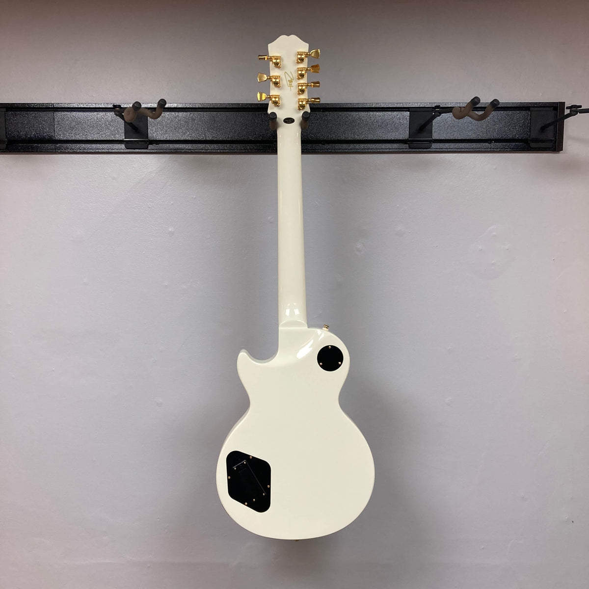 An Epiphone Matt Heafy Les Paul Custom Origins 7-string guitar in Bone White hangs on a black wall-mounted hanger, shown from the back with its gold tuning pegs visible.