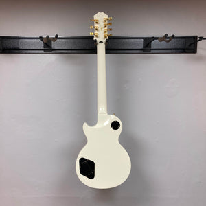 An Epiphone Matt Heafy Les Paul Custom Origins 7-string guitar in Bone White hangs on a black wall-mounted hanger, shown from the back with its gold tuning pegs visible.