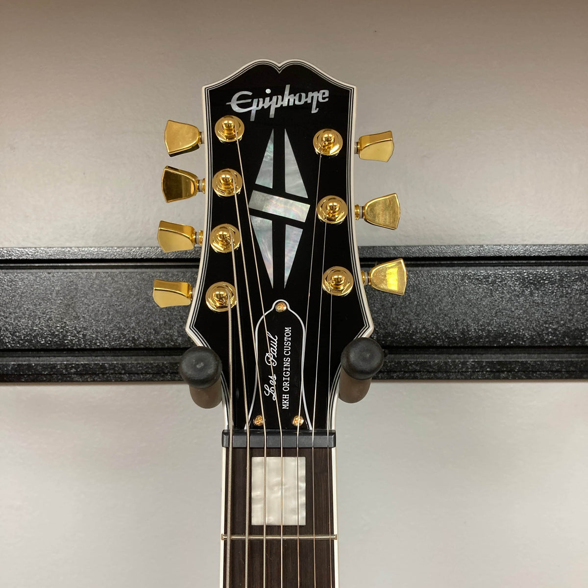 Close-up of an Epiphone Matt Heafy Les Paul Custom Origins, Bone White headstock with gold tuners, mother-of-pearl inlays, and Epiphone logo, mounted on a black wall hanger.