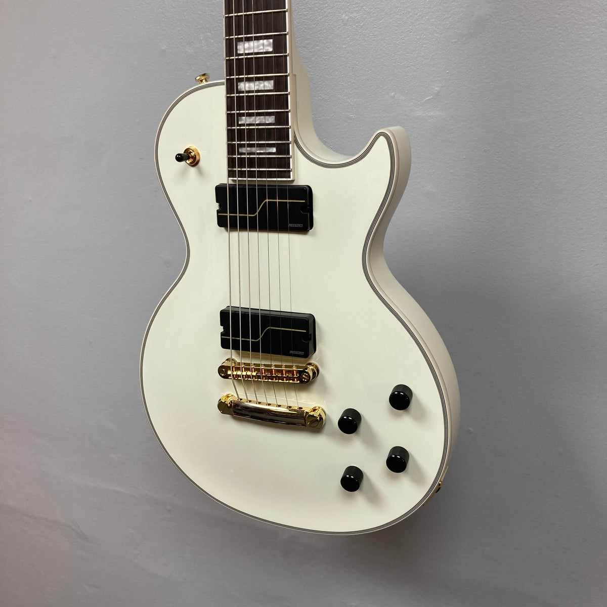 The Epiphone Matt Heafy Les Paul Custom Origins in Bone White, a 7-string guitar with gold hardware, two black pickups, three black knobs, and a switch, hangs on a light gray wall.