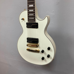 The Epiphone Matt Heafy Les Paul Custom Origins in Bone White, a 7-string guitar with gold hardware, two black pickups, three black knobs, and a switch, hangs on a light gray wall.