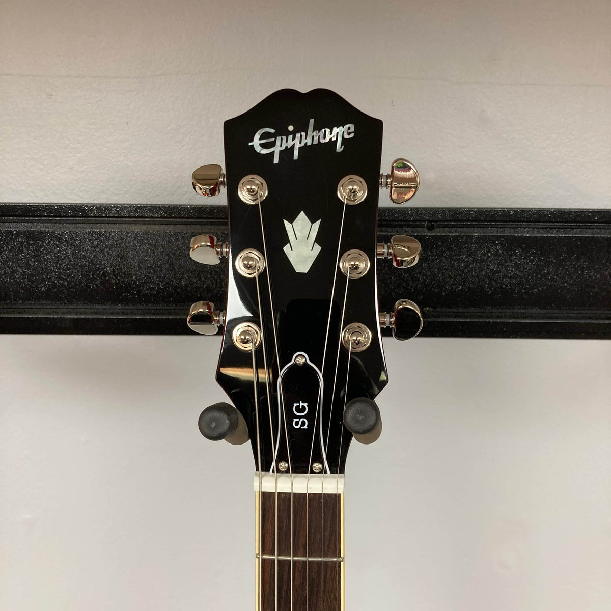 The Epiphone SG Standard Cherry w/Gigbag by Epiphone features a headstock with six tuning pegs, mounted on a black wall hanger. Inspired by the Gibson Collection, it has a mahogany body for classic rock style.