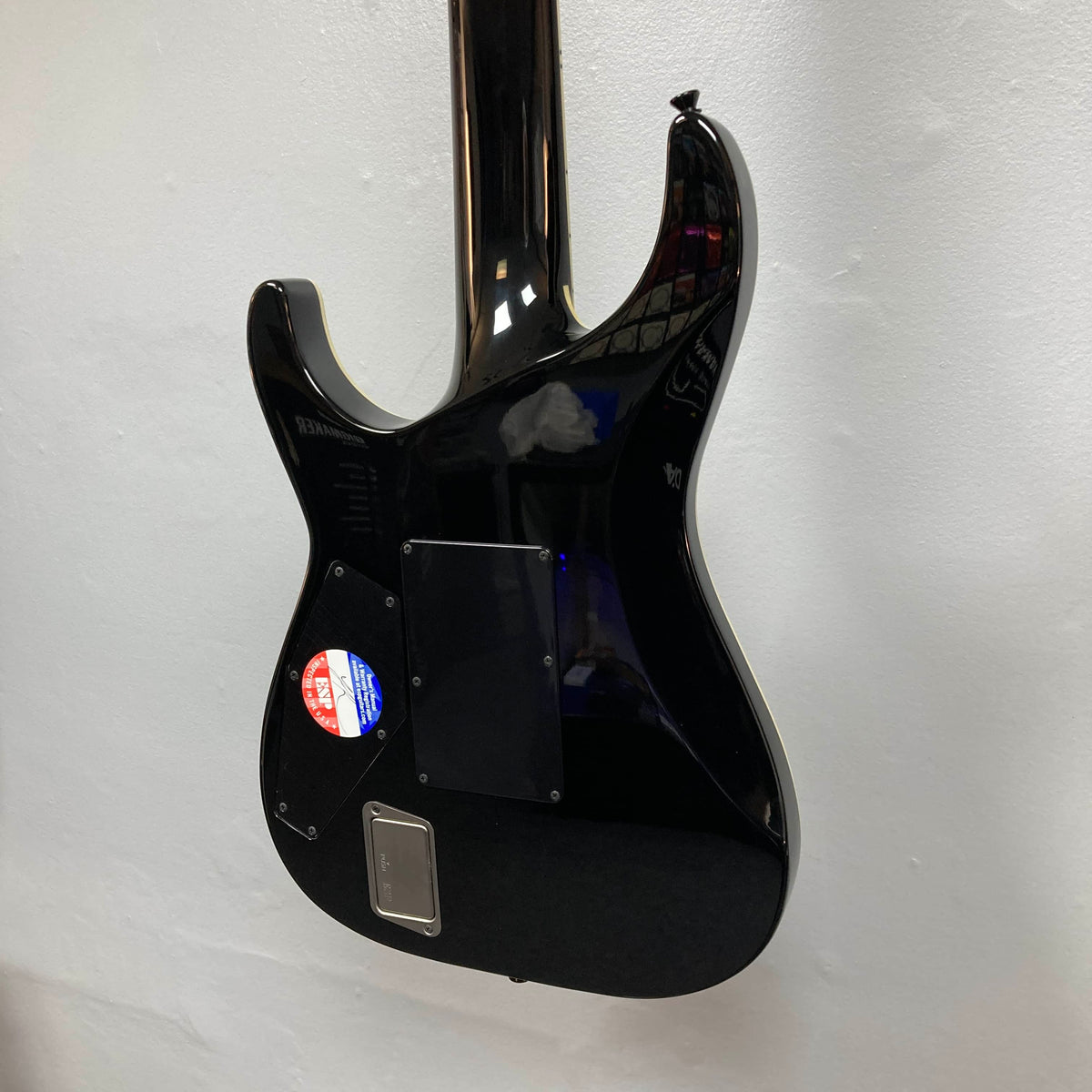 The back of the ESP E-II Horizon FR-7 Black 7-string electric guitar by ESP is shown against a white wall, displaying a sticker and several plastic covers over its electronics.