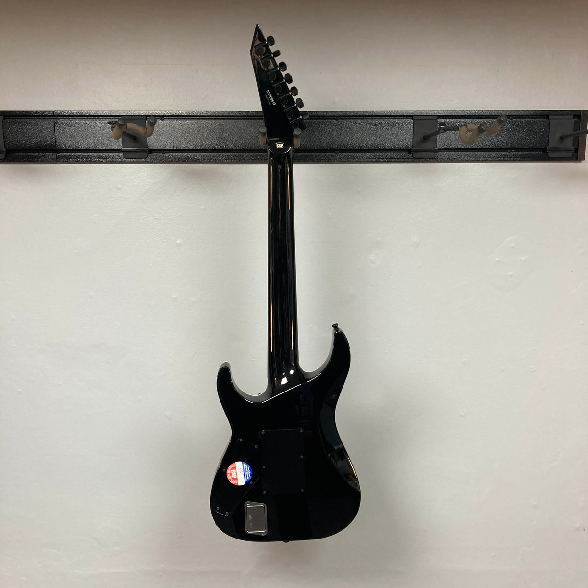 An ESP E-II Horizon FR-7 Black w/Case 7-string electric guitar by ESP hangs on a wall-mounted rack, displaying its back, a sticker near the bottom, and the distinctive Floyd Rose bridge.