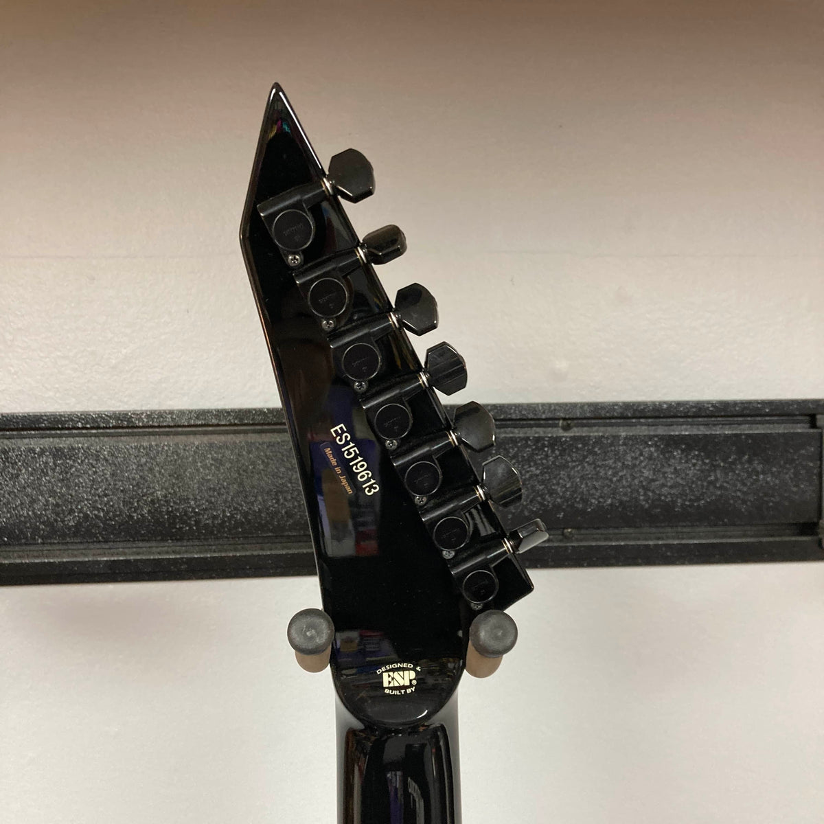 Close-up of the ESP E-II Horizon FR-7 Black electric guitar headstock, showing six tuning pegs and serial number ES176969, resting on a wall-mounted holder. Comes with case. Brand: ESP.