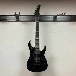An ESP E-II Horizon FR-7 Black w/Case 7-string electric guitar by ESP with a pointed headstock and Floyd Rose bridge hangs on a wall-mounted rack against a plain white wall.