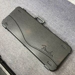 A black hard-shell Fender case with a curved design and molded handle, ideal for the Fender American Professional II Stratocaster HSS in Dark Night, rests on a gray carpeted floor.