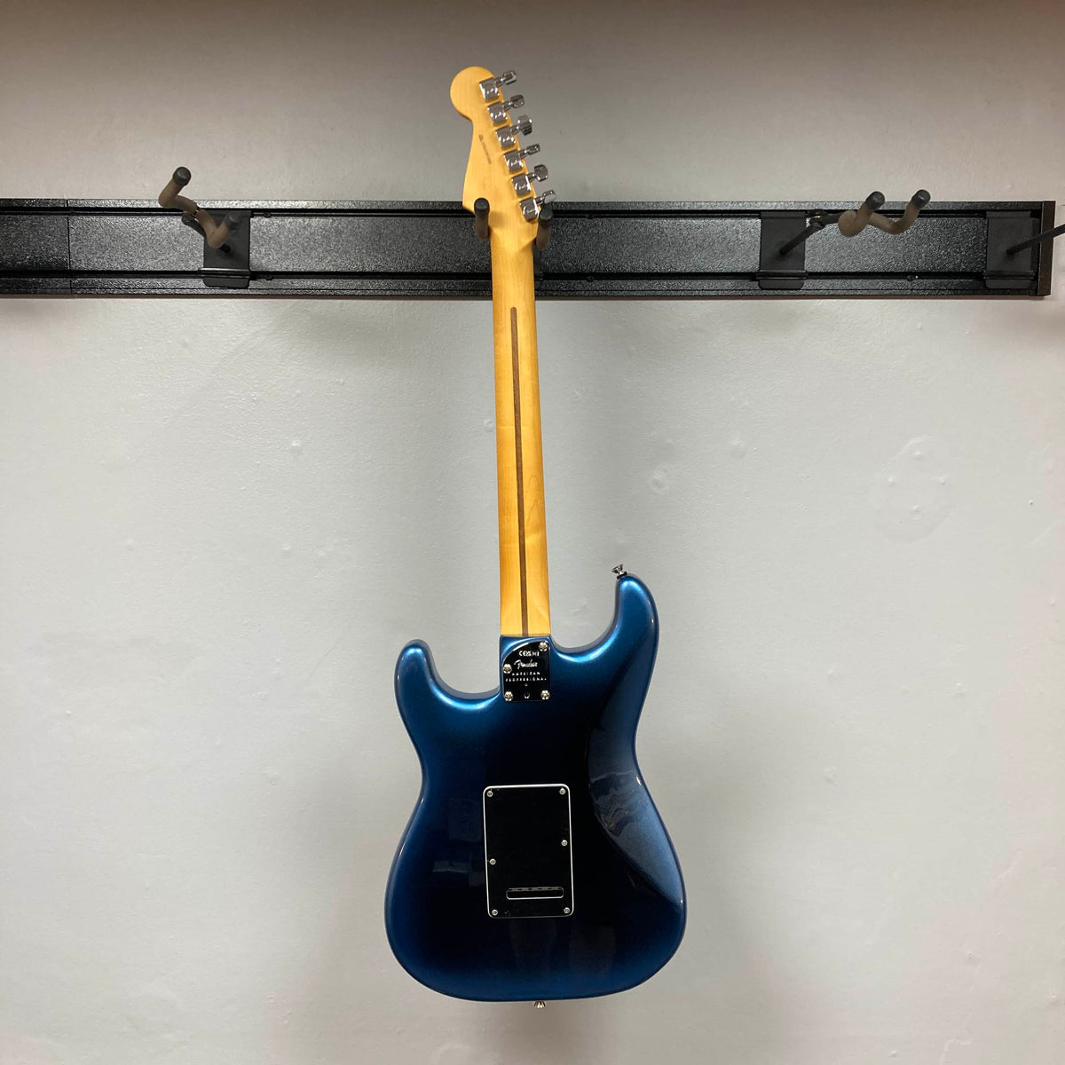 A Fender American Professional II Stratocaster HSS in Dark Night by Fender hangs by its neck on a wall-mounted rack, displayed from the back against a plain white wall.