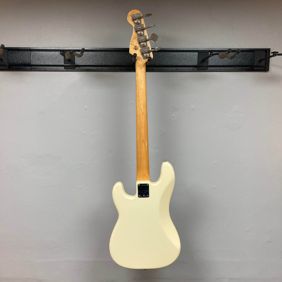 A 2005 Fender American Vintage '62 Precision Bass in Olympic White with natural wood neck hangs on a black wall-mounted rack, shown from the back against a plain gray wall. Includes case.