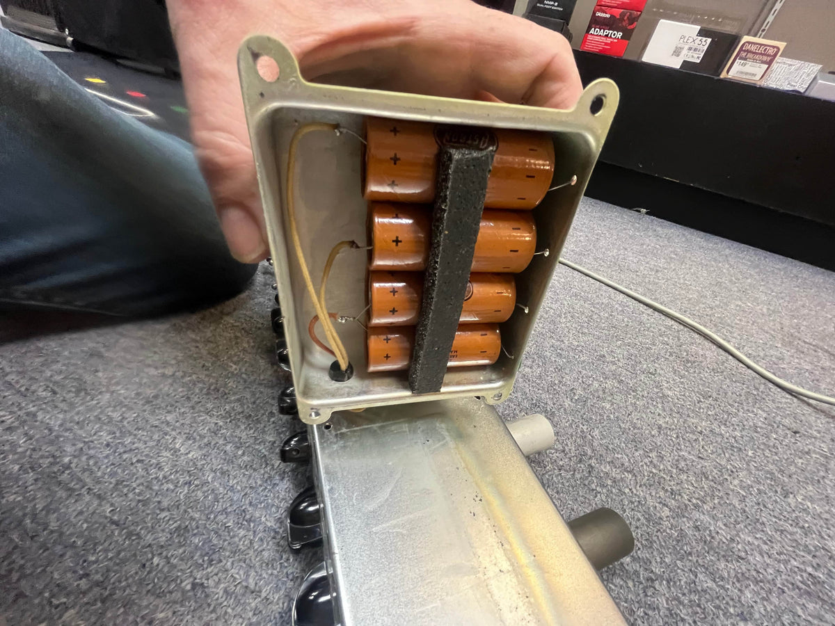 A hand opens a metal enclosure, revealing orange capacitors and wiring, resembling the iconic Fender 1957 Bassman 5F6 Narrow Panel 40-Watt 4x10" Guitar Combo w/Flight Case.