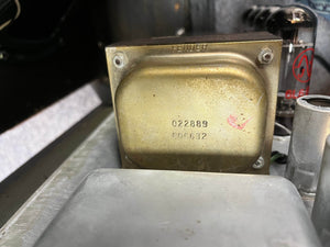 Close-up of a metallic part from a 1966 Fender Black Panel Twin Reverb 2-Channel 85-Watt 2x12" Guitar Combo Amp, engraved with "Fender" and stamped "022889" and "606632"; a vacuum tube appears in the background.