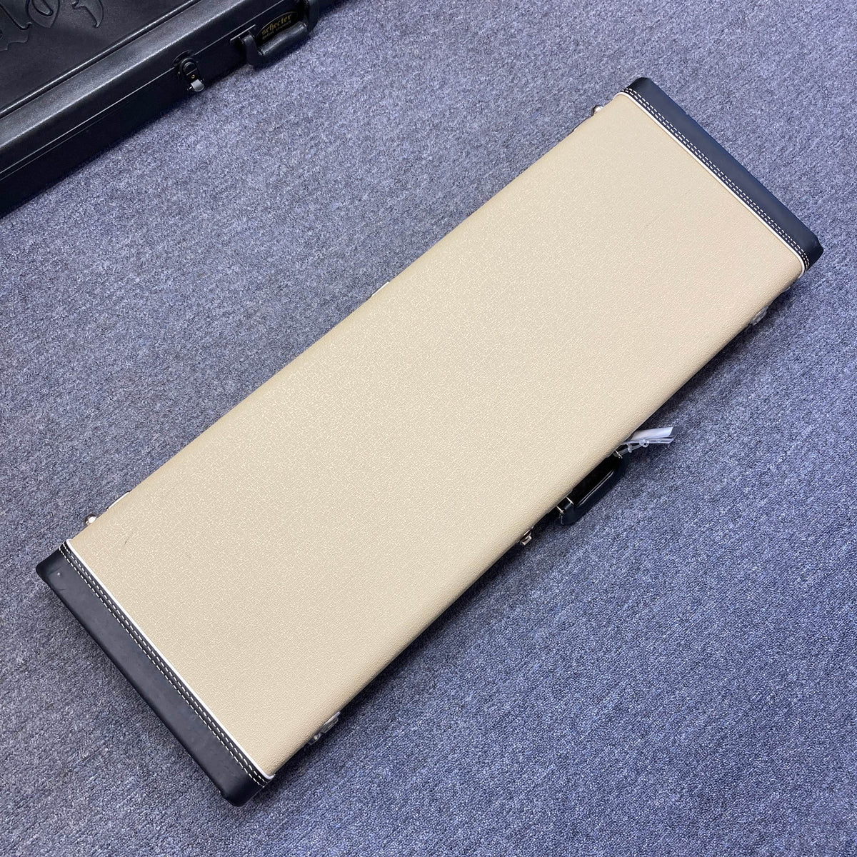 A rectangular beige case with black corners and edges sits closed on a gray carpeted floor, designed to protect the Fender 2006 Eric Johnson Stratocaster guitar.