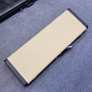 A rectangular beige case with black corners and edges sits closed on a gray carpeted floor, designed to protect the Fender 2006 Eric Johnson Stratocaster guitar.