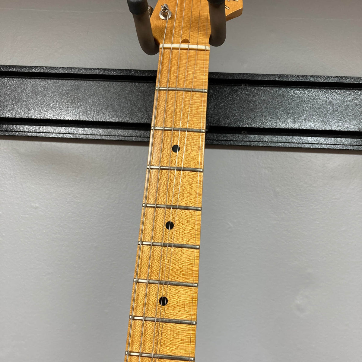 Close-up of a wooden neck with metal frets and dot inlays, hanging on a wall-mounted holder, featuring the iconic Fender 2006 Eric Johnson Stratocaster guitar's maple fingerboard.