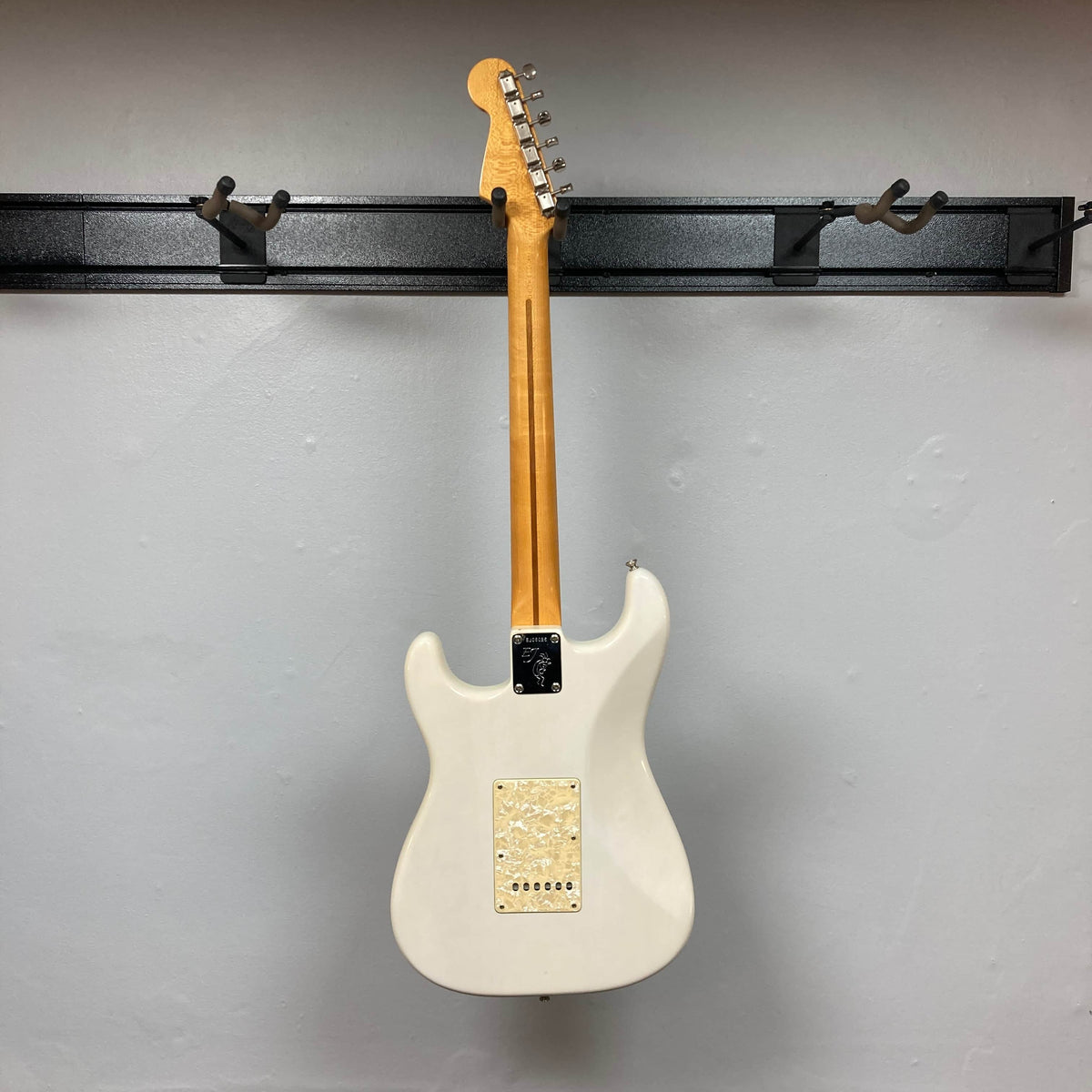 A 2006 Fender Eric Johnson Stratocaster electric guitar by Fender, white with a maple fingerboard, hangs by its neck on a black wall rack and is photographed from the back against a plain light gray wall.