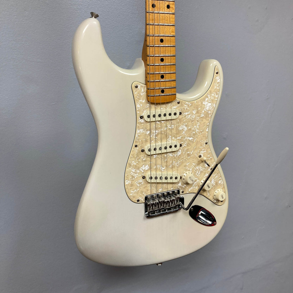 A 2006 Fender Eric Johnson Stratocaster by Fender, featuring a white finish, maple fingerboard, three single-coil pickups, white pearloid pickguard, and tremolo arm hangs on a gray wall.