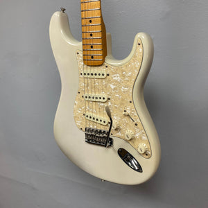 A 2006 Fender Eric Johnson Stratocaster in white, featuring a maple fingerboard, pearloid pickguard, three single-coil pickups, and a tremolo bridge, hangs against a gray wall.