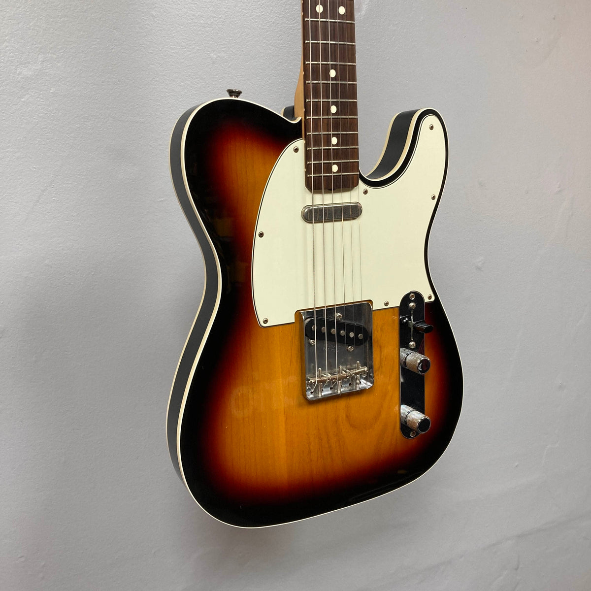 A Fender Heritage '60s Telecaster Custom Made in Japan Sunburst electric guitar with a white pickguard and two single-coil pickups hangs on a light gray wall, evoking vintage Telecaster style. Includes gigbag.