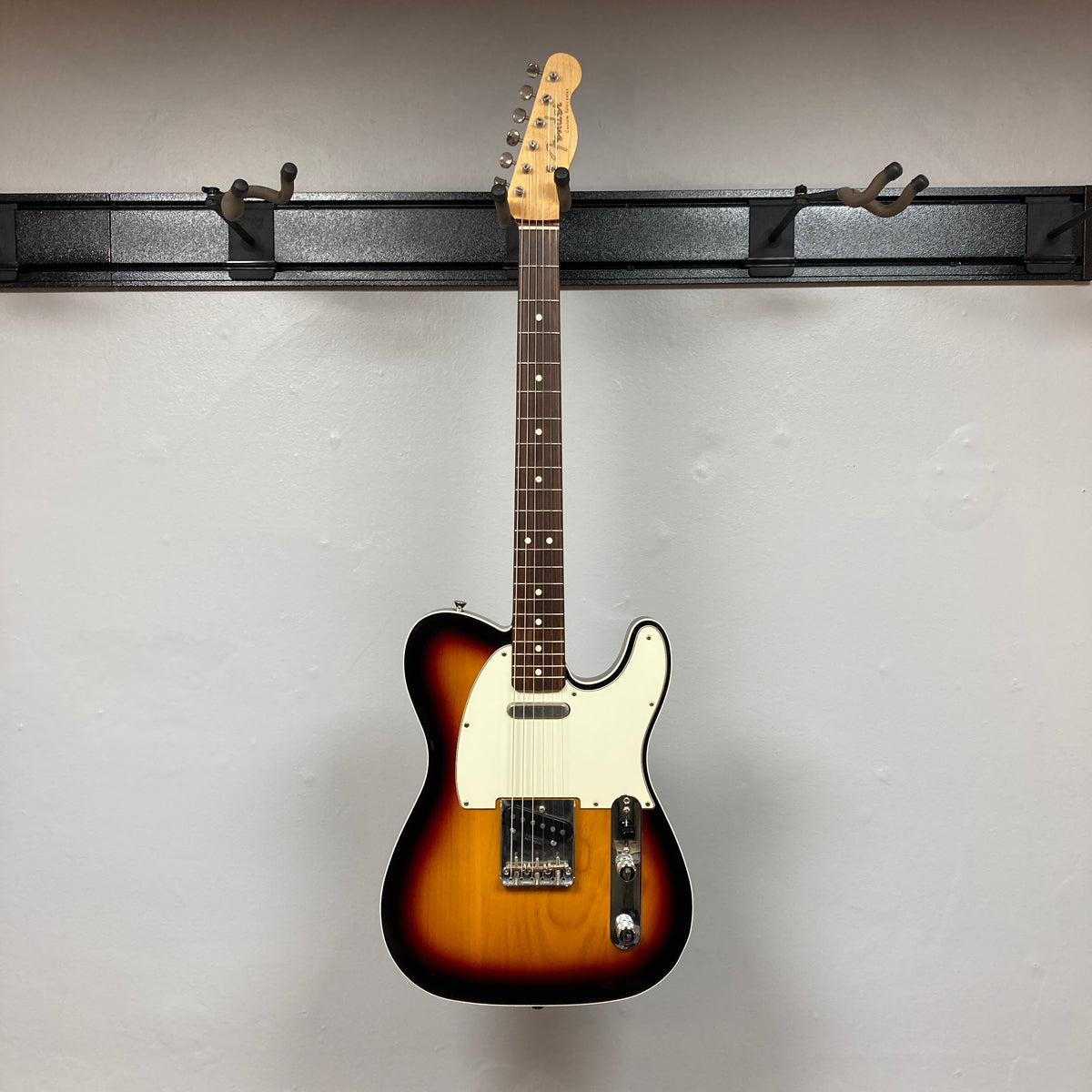 A Fender Heritage '60s Telecaster Custom Made in Japan Sunburst w/Gigbag by Fender hangs on a black wall-mounted guitar rack against a plain white wall.
