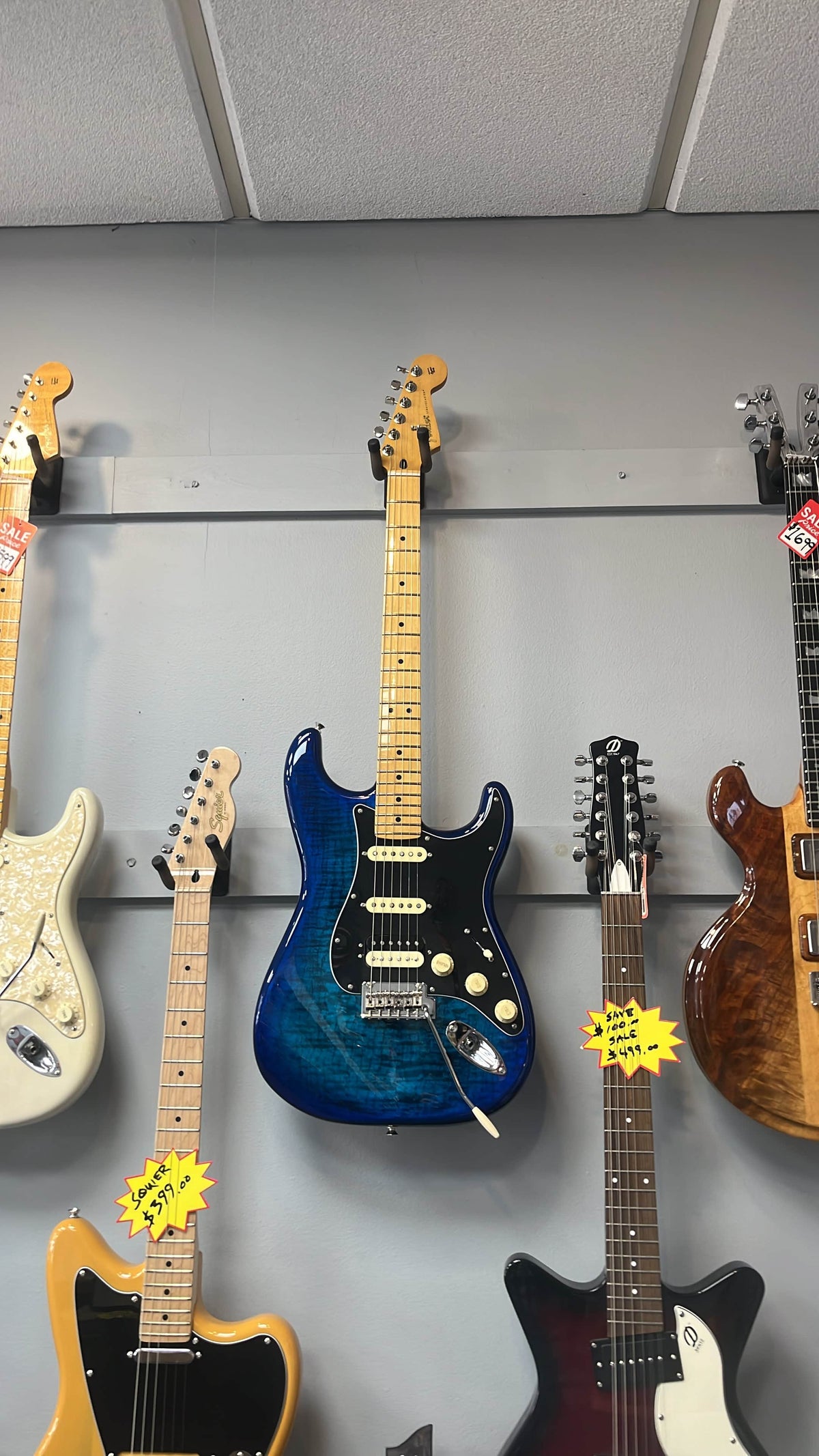 A used Fender Player II Stratocaster HSS Plus Top Maple Fingerboard Limited-Edition Electric Guitar in Blue Burst by Fender is shown on a wall with other guitars in a music store.