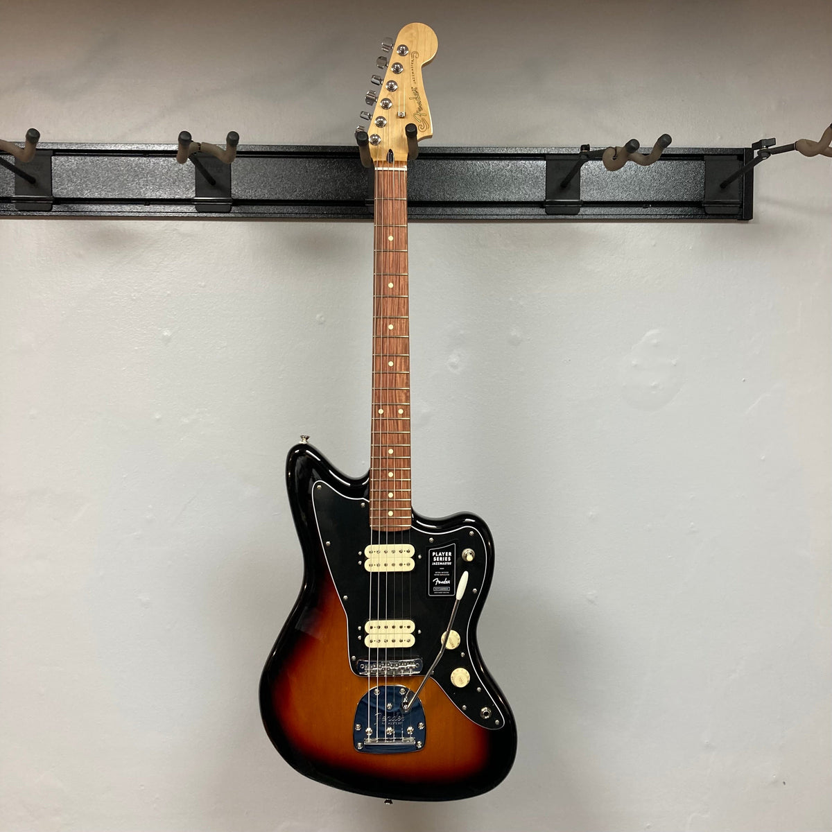 Fender Player Jazzmaster 3-Tone Sunburst Refurb w/Gigbag hanging on a wall hook, showcasing its sleek design and craftsmanship.