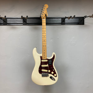 Fender Player Plus Stratocaster Olympic Pearl electric guitar on a wall hook, featuring black dot inlays and red and black hardware, with a gig bag included.