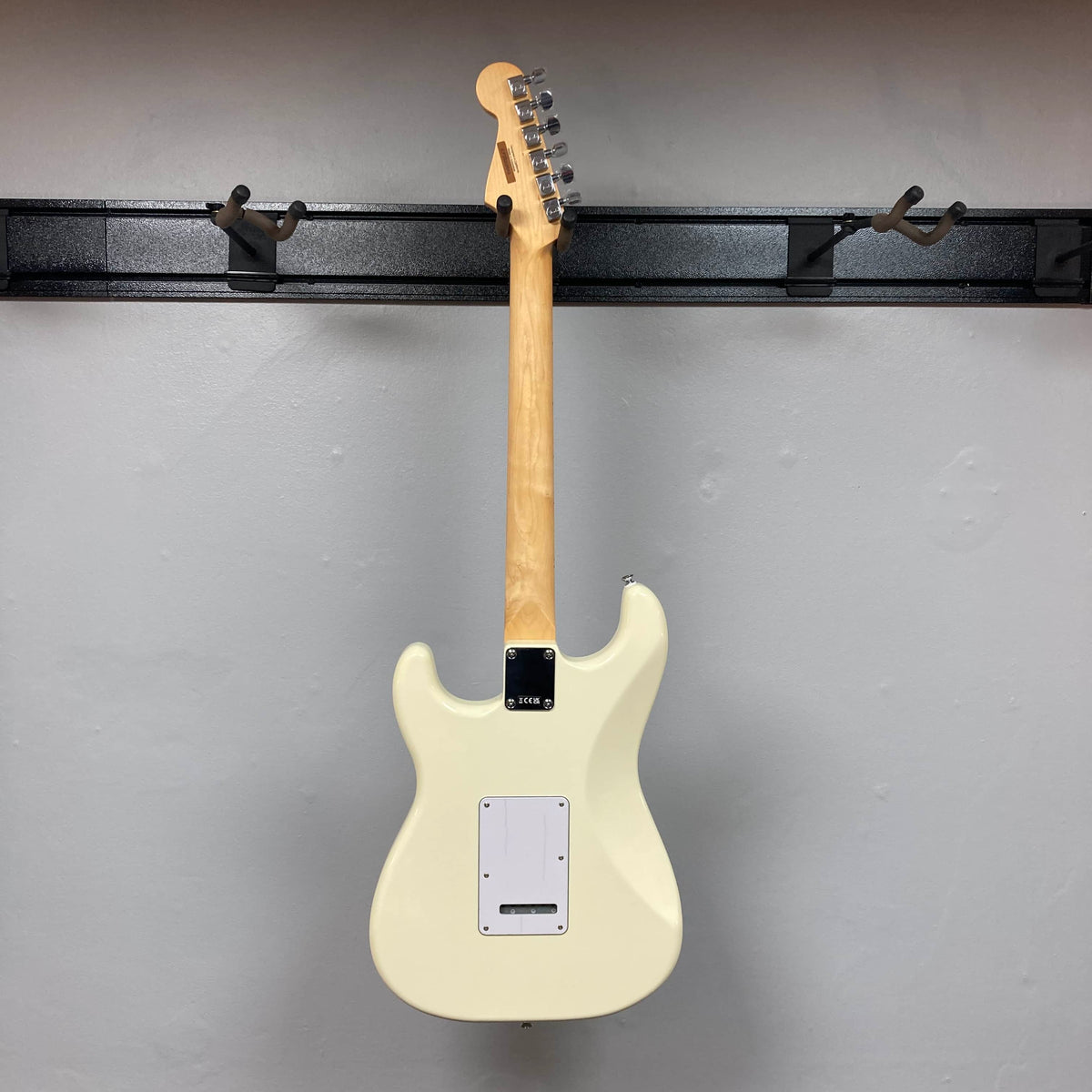A Fender Standard Stratocaster Olympic White Refurb by Fender hangs on a wall-mounted rack, shown from the back with its neck and tuning pegs clearly visible.