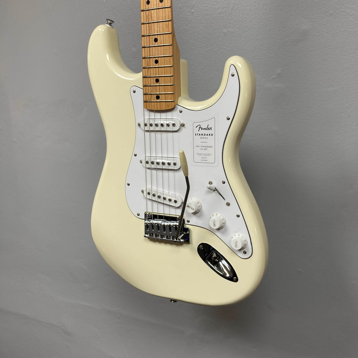 A Fender Standard Stratocaster Olympic White Refurb, featuring a maple neck and white pickguard, hangs against a plain gray wall.