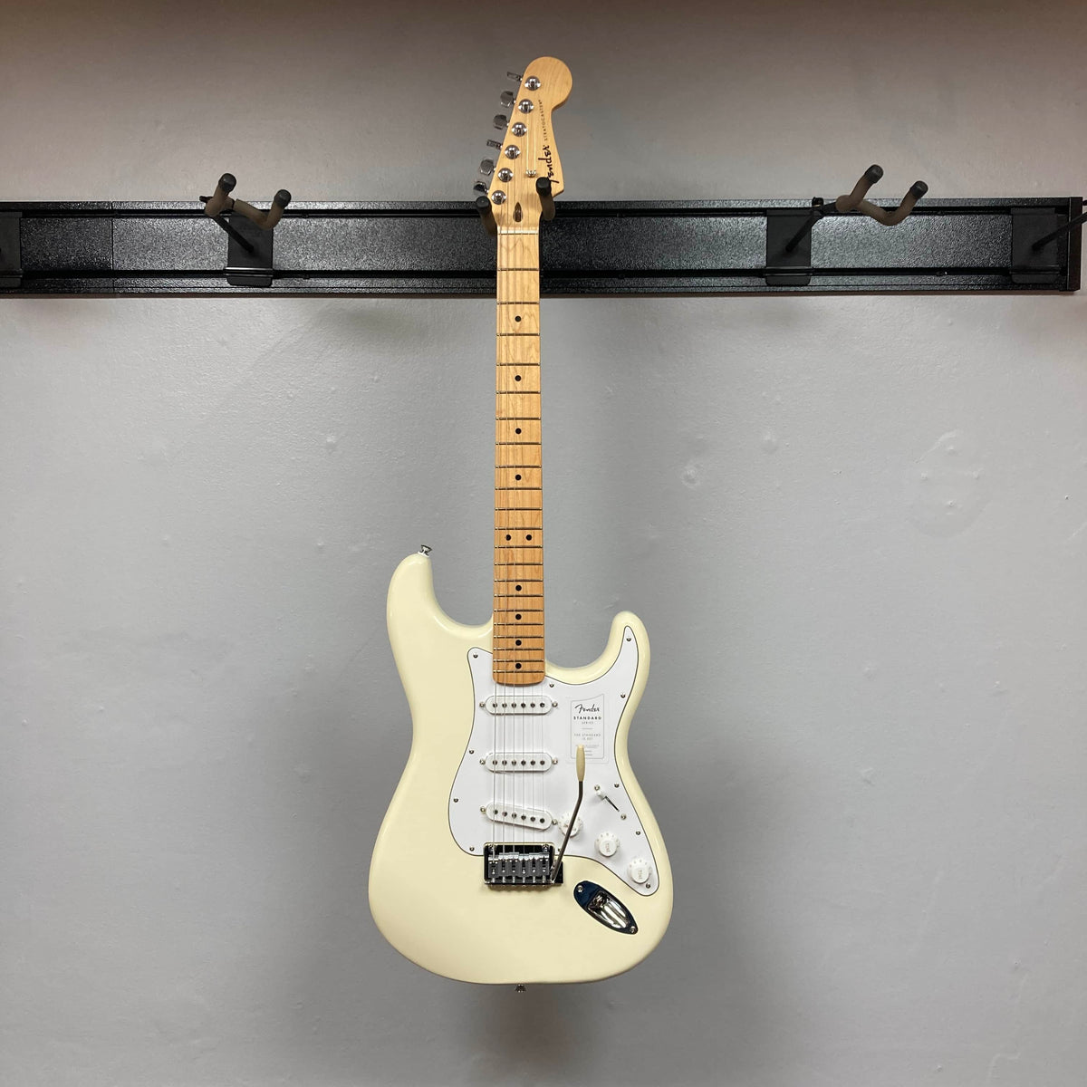 A Fender Standard Stratocaster Olympic White Refurb guitar by Fender hangs on a black wall-mounted rack against a light gray wall.