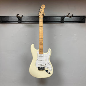 A Fender Standard Stratocaster Olympic White Refurb guitar by Fender hangs on a black wall-mounted rack against a light gray wall.