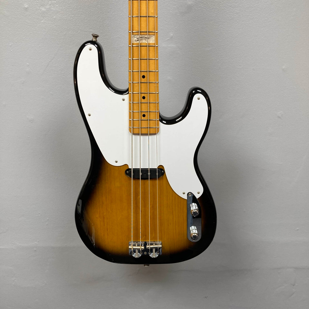A close-up of the Fender Sting P Bass Sunburst by Fender, featuring a vintage-style sunburst finish, white pickguard, and maple neck, photographed against a plain gray wall.